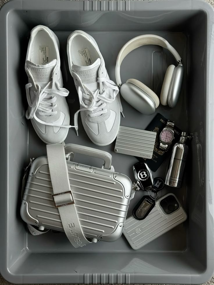 Curated grey travel bin with white sneakers, headphones, small luggage, and essentials